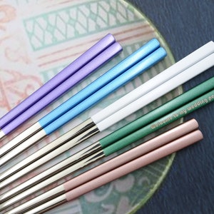 May include: Set of six pairs of chopsticks with different colored tips. The tips are purple, blue, white, green, and pink. The chopsticks are engraved with the text "Welcome to my wedding party - Sara".