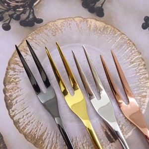 Op de afbeelding: Vier kleine vorken met verschillende afwerkingen: zwart, goud, zilver en rosé goud. De vorken zijn gerangschikt op een wit oppervlak met een decoratief bord op de achtergrond.