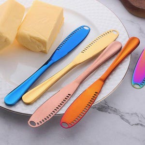 Puede incluir: Un conjunto de untadores de mantequilla de colores, azul, dorado, rosa y naranja, dispuestos sobre un plato blanco con mantequilla. Cada untador tiene una hoja ranurada y un mango redondeado. La mantequilla es de color amarillo claro.