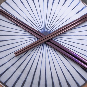 May include: A pair of purple metallic chopsticks crossed on a white and blue plate with a radial pattern.
