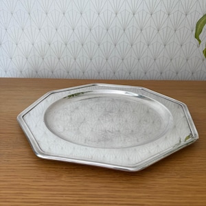 May include: A silver-coloured, octagonal serving tray with an oval centre. The tray has a reflective surface and a decorative border. The background features patterned wallpaper and a wooden surface.