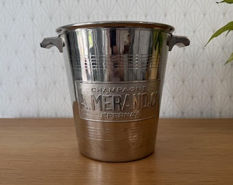 May include: A polished silver-tone metal champagne bucket featuring two side handles and a textured rectangular plaque on the front with the embossed text CHAMPAGNE A. MERAND & C EPERNAY. The bucket has horizontal grooved lines.