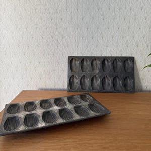 May include: Two vintage metal baking pans for making madeleines. The pans are gray and have a scalloped edge. The larger pan has 12 individual molds and the smaller pan has 18 individual molds.
