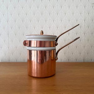 May include: A double boiler pot constructed from polished copper with a white ceramic insert. The pot features two long, angled copper handles extending from the sides and a matching copper lid with a small, rounded knob on the top center.