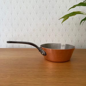 May include: A small copper saucepan with a dark metal handle. The interior of the pan is silver, contrasting with the copper exterior. The pan sits on a wooden surface, with a patterned wallpaper background.