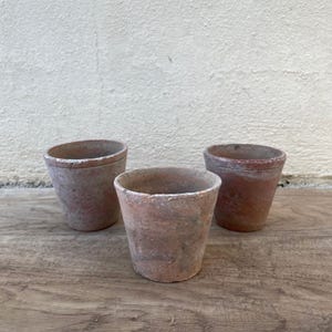 Op de afbeelding: Drie terracotta potten, elk met een iets andere bruine tint en een ruw, getextureerd oppervlak. De potten zijn gerangschikt op een houten oppervlak.