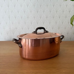 May include: A copper oval cooking pot with a lid and two side handles. The pot has a shiny, reflective surface and a dark handle on the lid. The pot is sitting on a wooden surface.