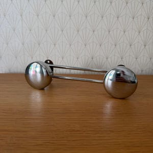 May include: A polished chrome cabinet handle featuring two spherical knobs connected by a double-bar bridge. The hardware has a reflective metallic finish and a horizontal orientation, designed for attachment to furniture or cabinetry doors and drawers.