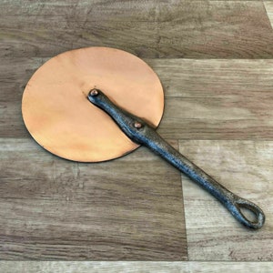 Puede incluir: Un cortador de pizza vintage de cobre con un mango largo de hierro negro. El mango tiene un bucle en el extremo para colgarlo.