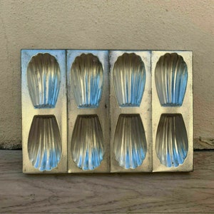 May include: A vintage metal baking pan with six individual molds for making madeleines. The molds are shaped like seashells and have a ribbed texture.