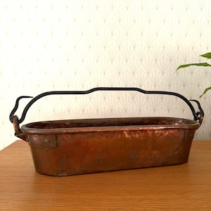 Puede incluir: Una jardinera rectangular de cobre con un asa de metal negro. La jardinera tiene una pátina oscura y envejecida y está remachada en las esquinas. El asa se arquea sobre la parte superior, sujeta a los lados. La jardinera está sobre una superficie de madera.