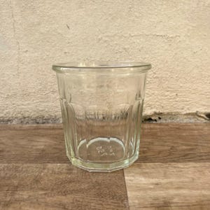Puede incluir: Un vaso transparente de estilo vintage con un borde grueso y redondeado y un cuerpo facetado. El vaso tiene una base ligeramente octogonal y está vacío. El vaso está sobre una superficie de madera.