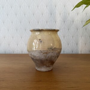 May include: A small, bulbous ceramic vase featuring a two-tone finish. The upper portion is coated in a smooth, glossy cream-coloured glaze, while the lower section displays a rough, textured, and weathered appearance in shades of grey and brown.