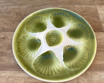 May include: A white ceramic oyster plate with six green shell-shaped indentations. The plate has a green rim and a white center.