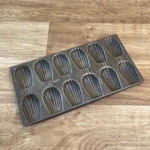 May include: A vintage metal baking pan with 12 individual moulds shaped like seashells. The pan is made of dark grey metal and has a slightly worn appearance.