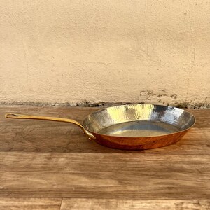 Puede incluir: Una sartén ovalada de cobre martillado con un mango de latón. La sartén está sobre una superficie de madera.