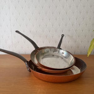 Op de afbeelding: Drie koperen pannen met zwarte handvatten zijn op een houten oppervlak gestapeld. De pannen variëren in grootte, met de grootste onderaan en de kleinste bovenaan. Het koper heeft een glanzend, reflecterend oppervlak.
