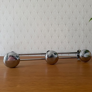 Peut inclure: Un porte-manteau mural horizontal composé de trois crochets sphériques en chrome poli fixés sur une structure métallique à deux barres. Le design comprend des plaques de fixation circulaires à chaque extrémité pour suspendre des vêtements.