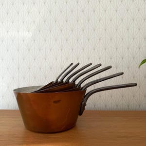 May include: A set of copper saucepans with dark metal handles. The pans are nested inside each other, with the largest one in the front. The copper has a warm, reddish-brown color, and the handles are curved.