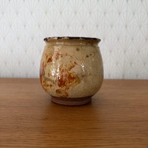 Puede incluir: Una pequeña vasija de cerámica redondeada con un esmalte moteado en beige y tostado que presenta parches irregulares de color naranja quemado y marrón. El borde es oscuro y texturizado, mientras que la base es de arcilla rojiza sin esmaltar.