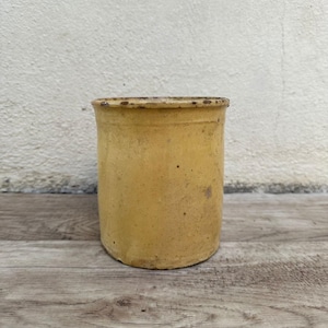 May include: A yellow ceramic pot with a worn, rustic finish. The pot is cylindrical in shape and has a slightly rounded bottom.