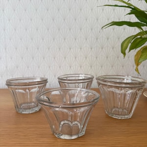 May include: Four clear glass dessert bowls with a heart-shaped design at the base. The bowls have a flared shape and are on a wooden surface. The background features patterned wallpaper and a green plant.