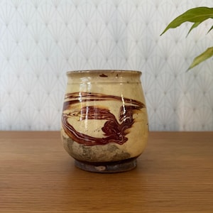 Puede incluir: Una taza de cerámica redondeada con una base de color crema y patrones de mármol en marrón oscuro. El recipiente tiene un acabado liso y brillante en la parte superior y una base gris mate texturizada, mostrando un contraste de color.