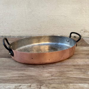 May include: An oval copper baking dish with two black handles. The dish has a worn, aged look and is sitting on a wooden surface.