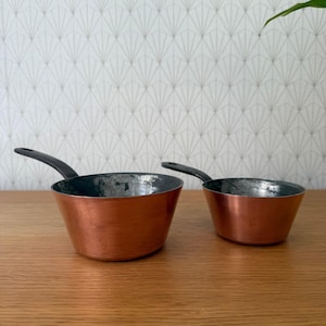 May include: Two copper saucepans with black handles. The pans have a shiny copper exterior and a dark interior. They are sitting on a wooden surface, with a patterned wallpaper background.