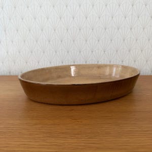 May include: An oval-shaped ceramic baking dish with a smooth, light tan interior and a darker, earthy brown exterior. The dish features a shallow depth and a simple, unadorned rim, showcasing a natural, glazed finish throughout the entire piece.