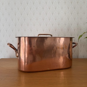 May include: A rectangular copper roasting pan with rounded corners, featuring two side handles and a matching lid with a central handle. The polished metal surface reflects light, showing a warm, metallic orange-brown hue throughout.