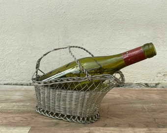 Puede incluir: Porta botellas de vino de alambre plateado con una botella de vino de vidrio verde en su interior. El porta botellas tiene un asa y tiene forma de cesta.