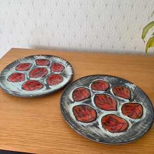 May include: Two oval ceramic plates with a black rim and a light blue interior. Each plate features seven red leaf-shaped designs with black detailing. The plates are on a wooden surface.