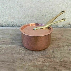 May include: A small copper saucepan with a brass handle and lid. The saucepan is sitting on a wooden surface.