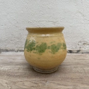 May include: A small, round, yellow ceramic pot with a green glaze design. The pot has a slightly rounded bottom and a wide opening.
