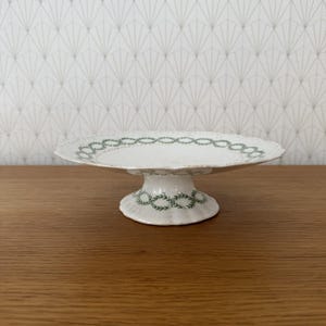 Può includere: Un'alzata per dolci in ceramica bianca con bordo smerlato e un motivo a ghirlanda di foglie di alloro verdi che circonda il bordo e la base. Il supporto presenta un piatto largo e poco profondo sostenuto da una base robusta e svasata.