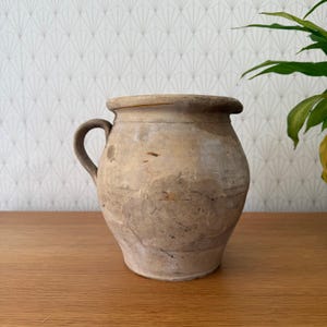 Może przedstawiać: Rustykalny dzbanek ceramiczny w stylu antycznym z jednym uchwytem. Dzbanek ma szeroki, zaokrąglony korpus i lekko rozszerzoną krawędź. Zewnętrzna strona ma nakrapiany beżowy kolor z ciemniejszymi brązowymi i czarnymi oznaczeniami.