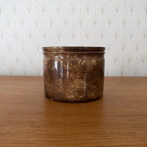 May include: A cylindrical ceramic jar featuring a mottled brown and tan glaze with a speckled texture. The vessel has a smooth, straight-sided body and a slightly raised rim at the top, showcasing a glossy finish throughout the exterior surface.