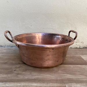 May include: A copper bowl with two curved handles. The bowl has a wide, shallow design and a polished finish. The copper has a warm, reddish-brown hue, and the bowl appears to be vintage or antique.