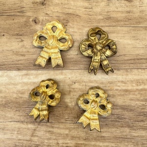Könnte beinhalten: Vier goldfarbene, dekorative Schleifen aus Metall mit einem erhabenen Design. Die Schleifen sind in einem quadratischen Muster auf einer Holzoberfläche angeordnet.