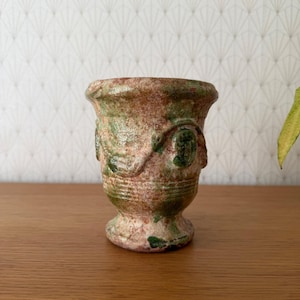 May include: A small, antique-style vase with a pedestal base. The vase is primarily brown with green and red accents, and has a textured surface. Decorative elements are visible on the sides. The vase is likely made of ceramic or pottery.