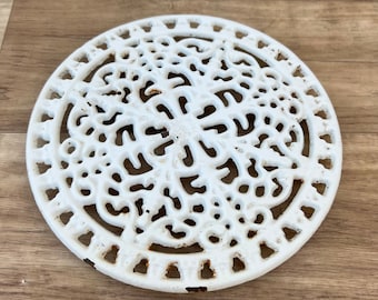 May include: White cast iron trivet with a decorative, intricate design. The trivet is round and has a scalloped edge.