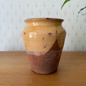 Puede incluir: Una maceta de cerámica rústica de dos tonos. La parte superior es de un amarillo cálido, mientras que la mitad inferior es de un marrón rojizo oscuro. La maceta tiene una abertura ancha y una forma ligeramente cónica. Está sobre una superficie de madera.