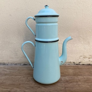 May include: A vintage light blue enamel coffee pot with black trim. The pot features a cylindrical upper section and a conical lower section, with a curved spout and two loop handles. The lid is also light blue with a black trim.