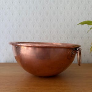 May include: A copper bowl with a rounded shape and a single ring handle. The bowl has a polished finish, reflecting the light. The bowl is sitting on a wooden surface.