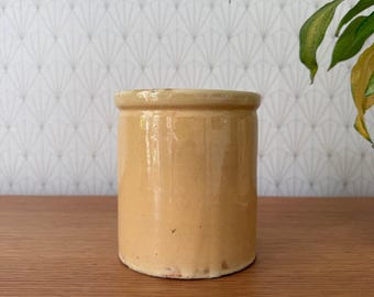 May include: A cylindrical, antique, tan-colored ceramic jar with a slightly uneven rim. The jar has a smooth, glossy finish and sits on a wooden surface. The background features a patterned wallpaper and a glimpse of a green plant.