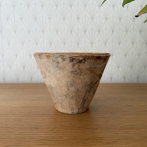May include: A small, cone-shaped, handmade pottery vessel. The exterior is a mottled blend of tan, brown, and gray, with a rough, textured surface. The rim is a darker shade of brown. The vessel sits on a wooden surface.
