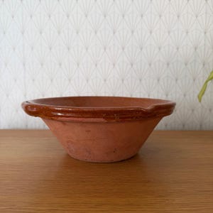 May include: A shallow, round terracotta bowl with a wide, flat rim. The exterior and base are unglazed, showing a natural matte reddish-brown clay texture, while the top rim features a smooth, glossy, dark brown glazed finish for contrast.