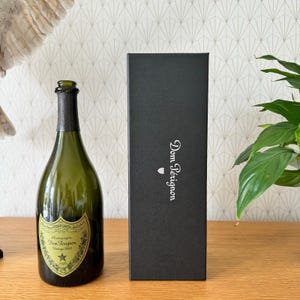 Könnte beinhalten: Eine dunkelgrüne Glasflasche Dom Pérignon Vintage 2000 Champagner mit einem cremefarbenen Etikett mit schwarzem Text und einem Sternsymbol, neben einer hohen, rechteckigen schwarzen Geschenkbox mit dem vertikal aufgedruckten weißen Text Dom Pérignon.