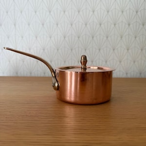 May include: A small copper saucepan with a long, curved handle and a lid. The pot has a shiny, reflective surface and a brass-coloured handle. The background is a light-coloured wall with a repeating geometric pattern. The saucepan is on a wooden surface.